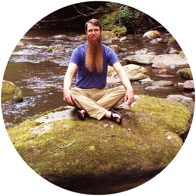 thomoas ealey seated in lotus pose atop a moss-covered rock with a river behind him